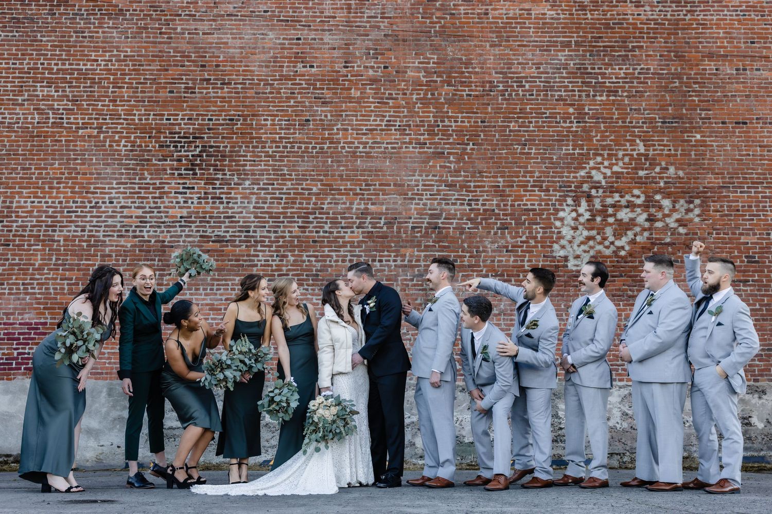 Wedding party poses against brick wall with groomsmen in light suits and bridesmaids in dark dresses.