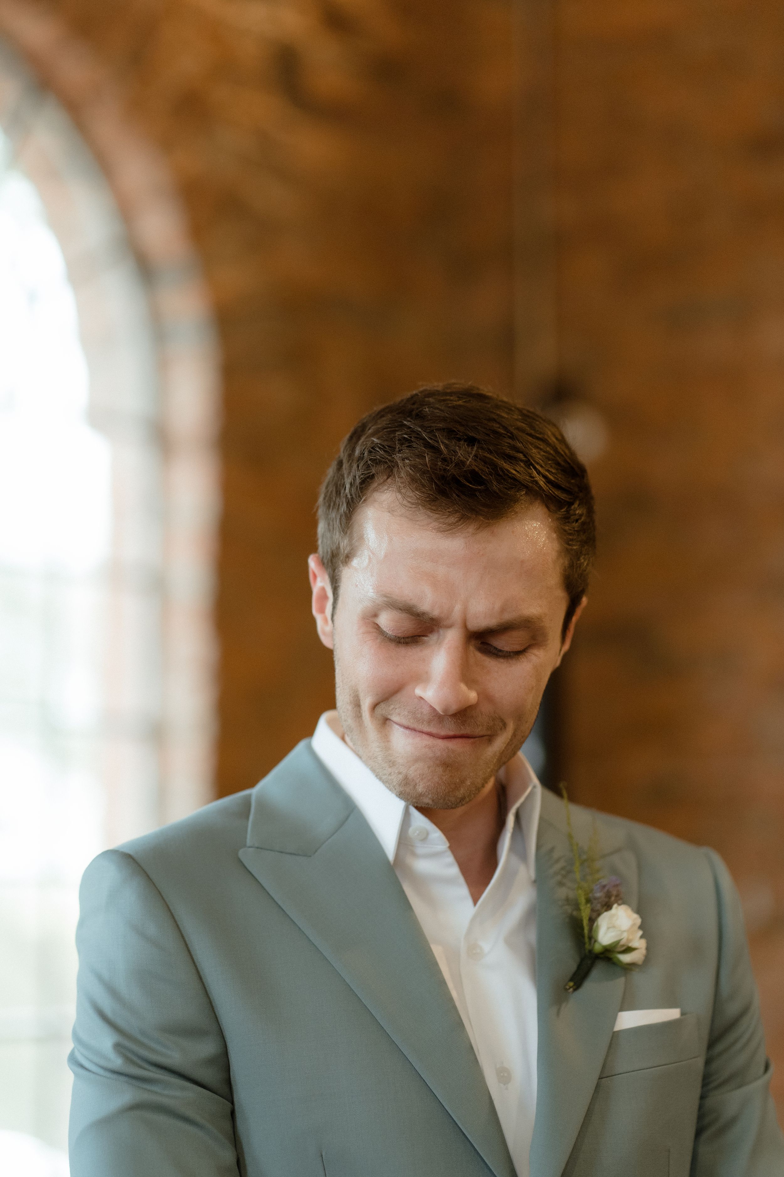 A groom in a light gray suit jacket and white shirt smiles while looking down during a wedding ceremony.