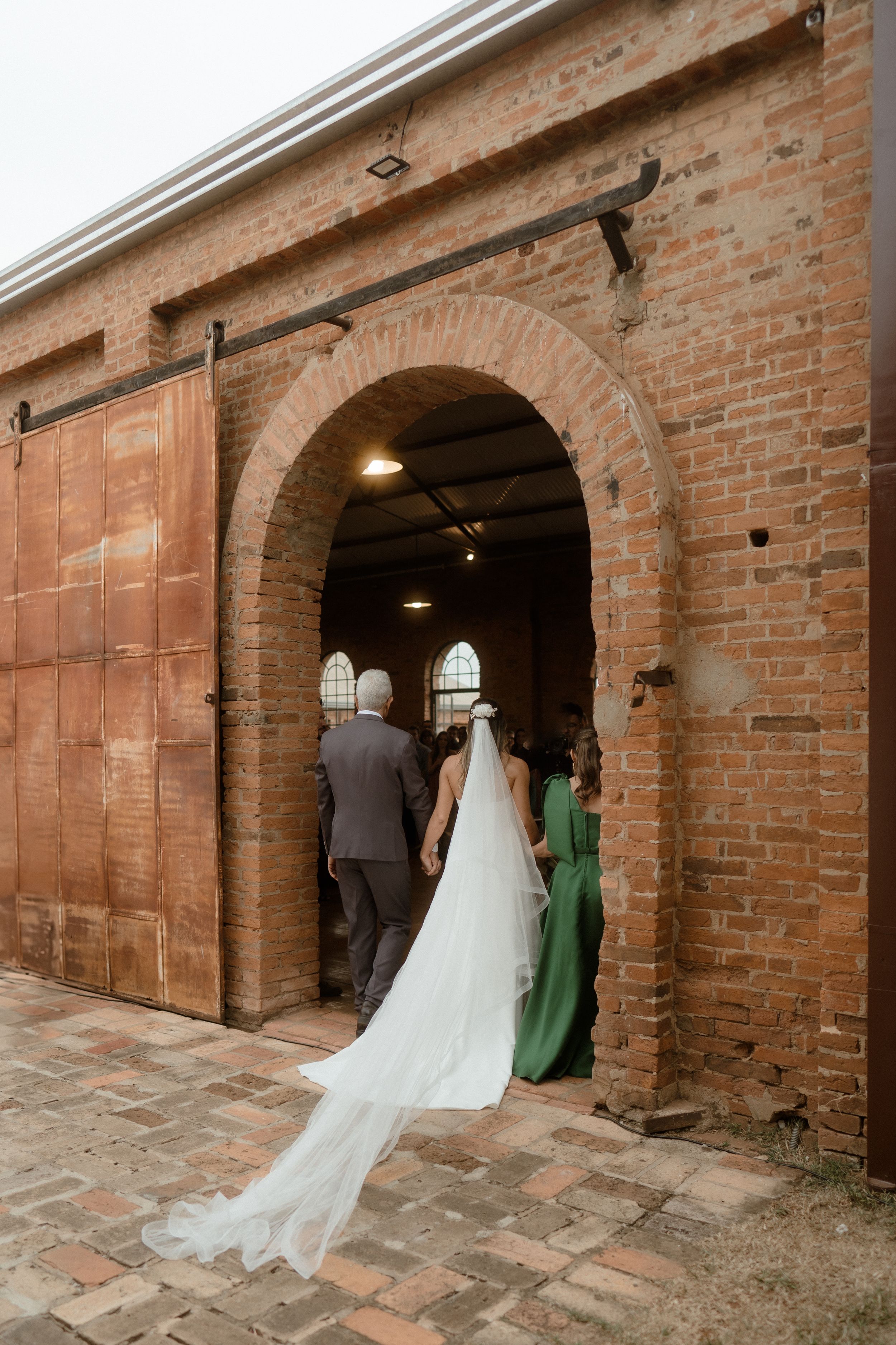 Wedding party enters through rustic brick archway with flowing white bridal gown train visible.