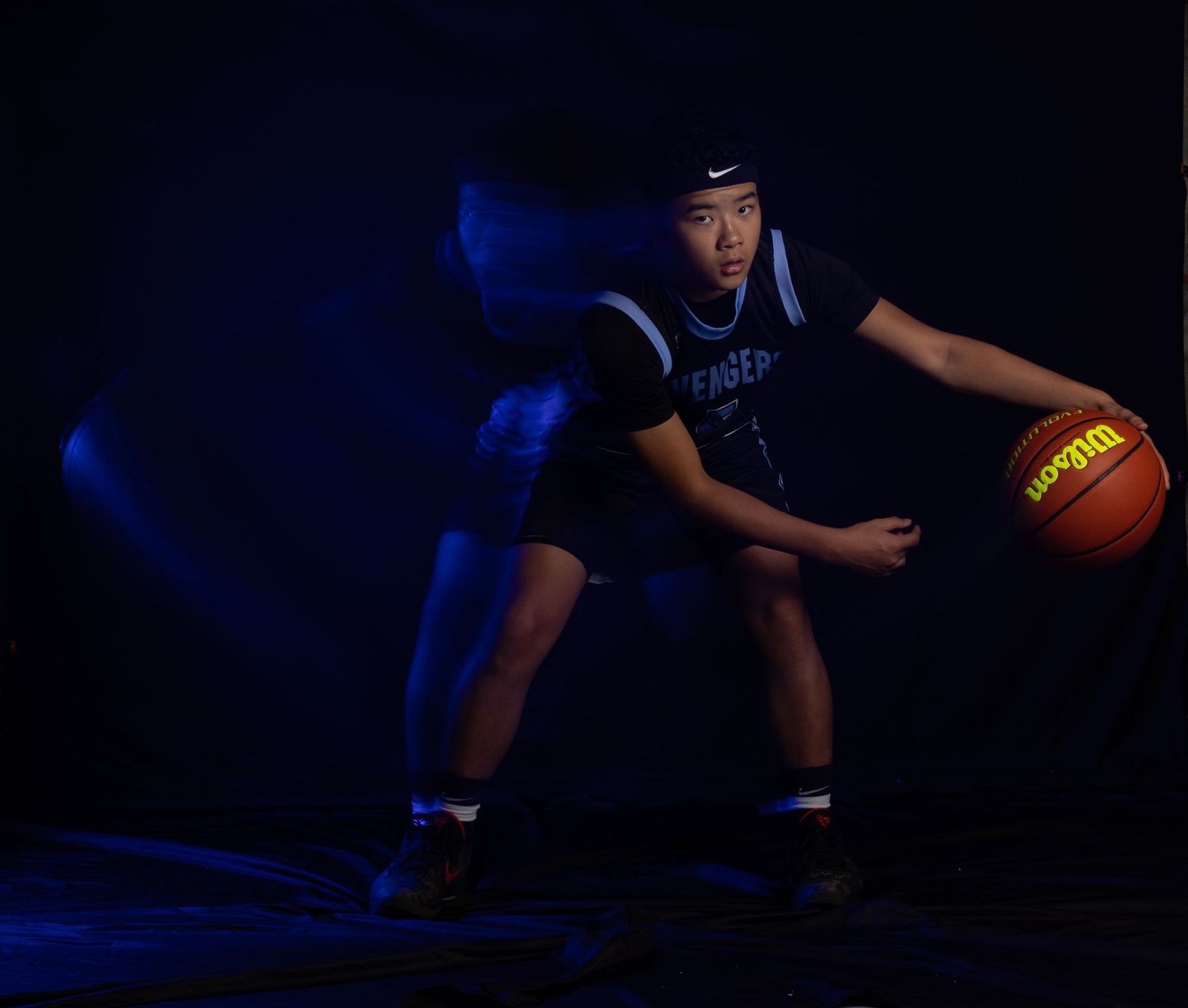 Basketball player crouches in defensive stance with basketball against dark blue studio lighting.