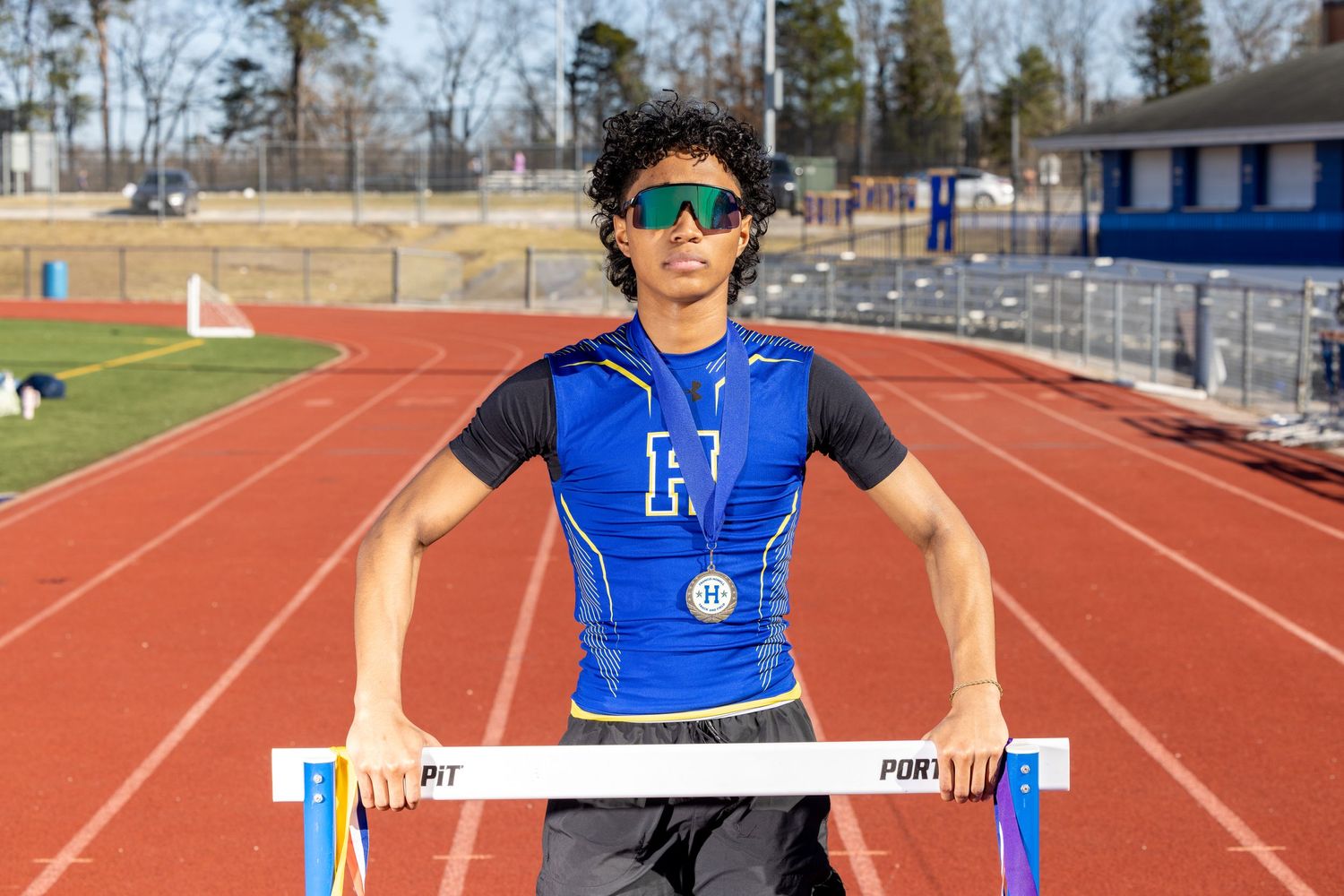 An athlete in a blue shirt and sunglasses stands behind a track hurdle on a red running track.