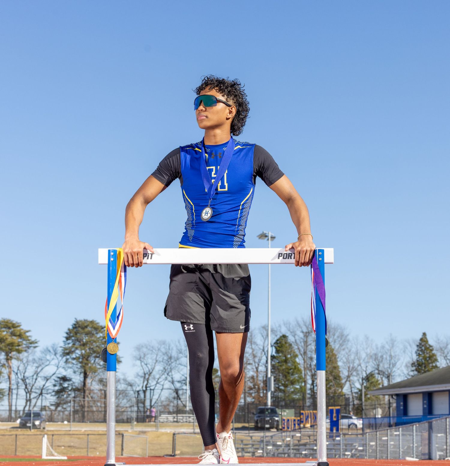 Track athlete in blue uniform stands confidently on a hurdle against clear blue sky at outdoor stadium track.