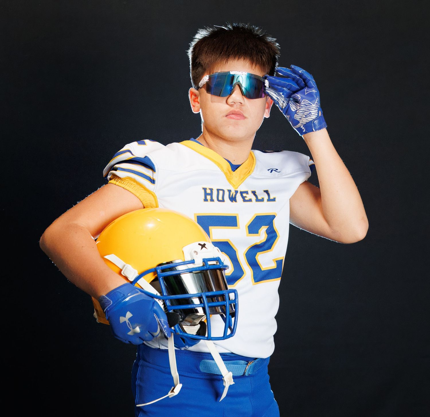 Football player in white and blue uniform holds helmet and adjusts sunglasses while posing against dark background.