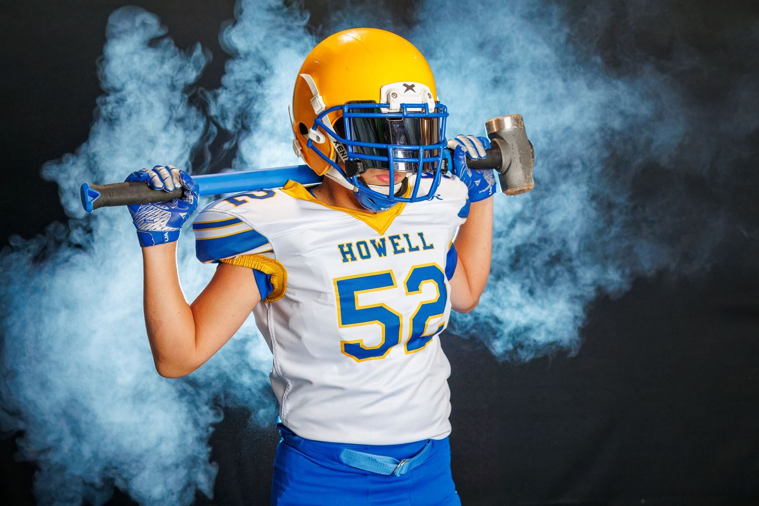 High school football player in white and blue Howell jersey number 52 poses dramatically with smoke effect against dark background.