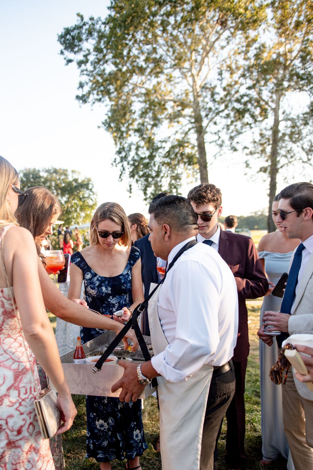 A group of formally dressed guests gather outdoors at a sunny summer wedding around the oyster shucker at cocktail hour.