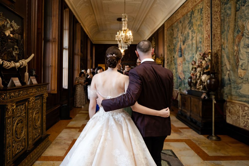A couple in wedding attire embrace while overlooking an elegant hallway with tapestries adorning the walls of the historic mansion.