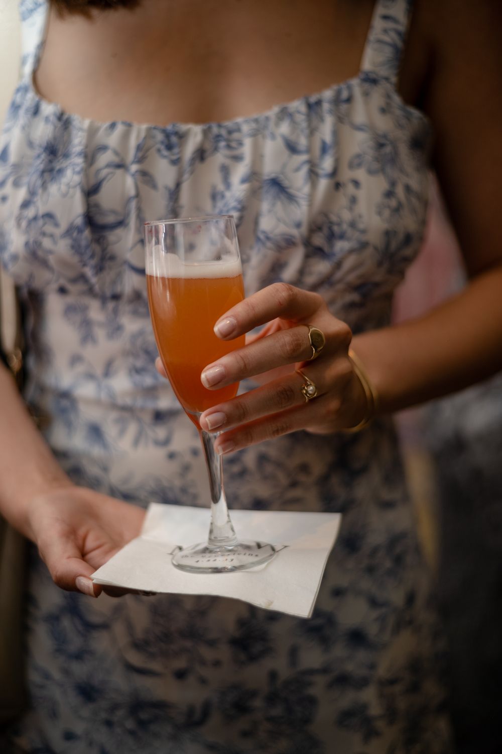 A floral dress-clad woman holds the specialty cocktail in a champagne glass on a white napkin.