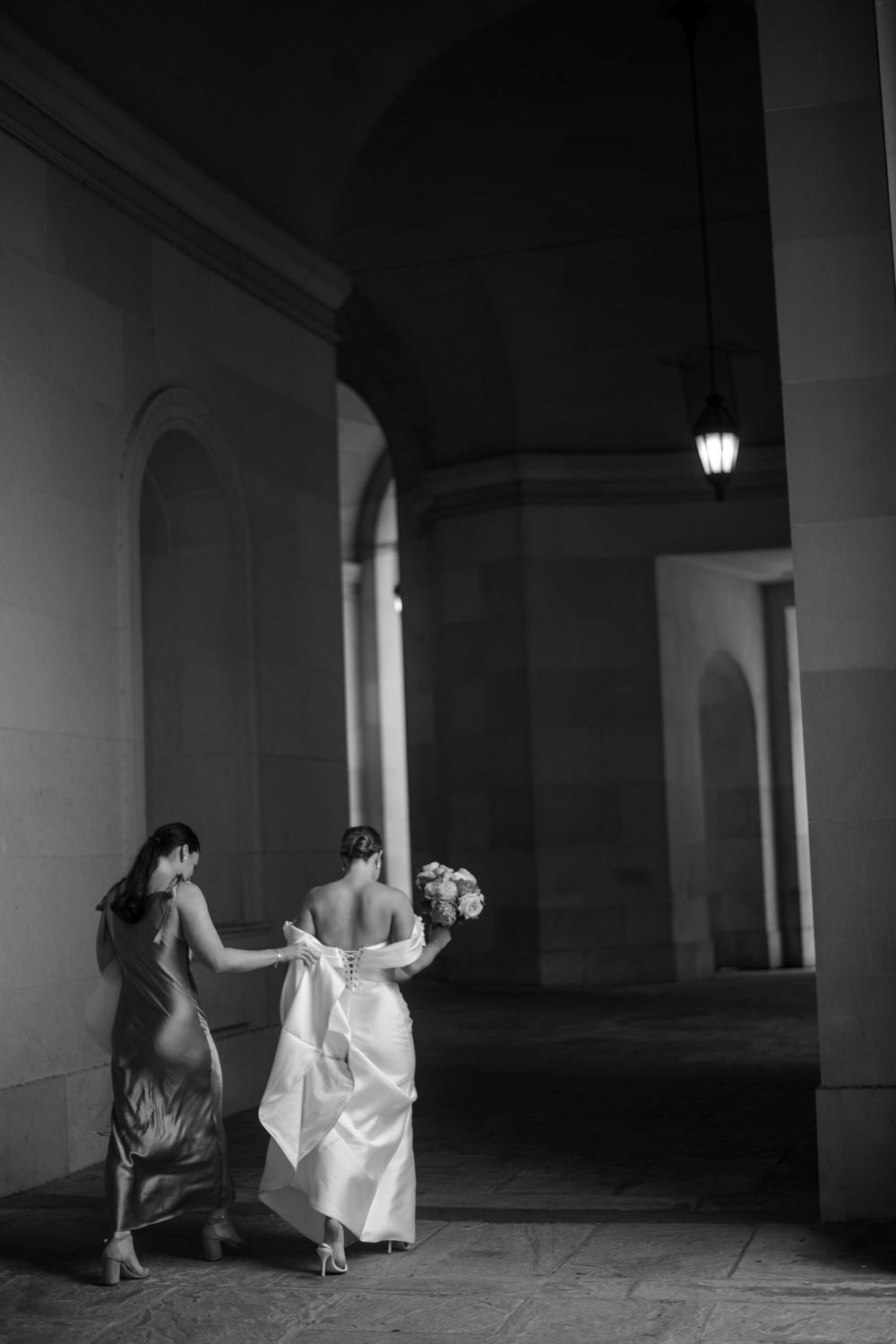 Bride being walked through an arched corridor in a dramatic black and white scene with her bridesmaid.