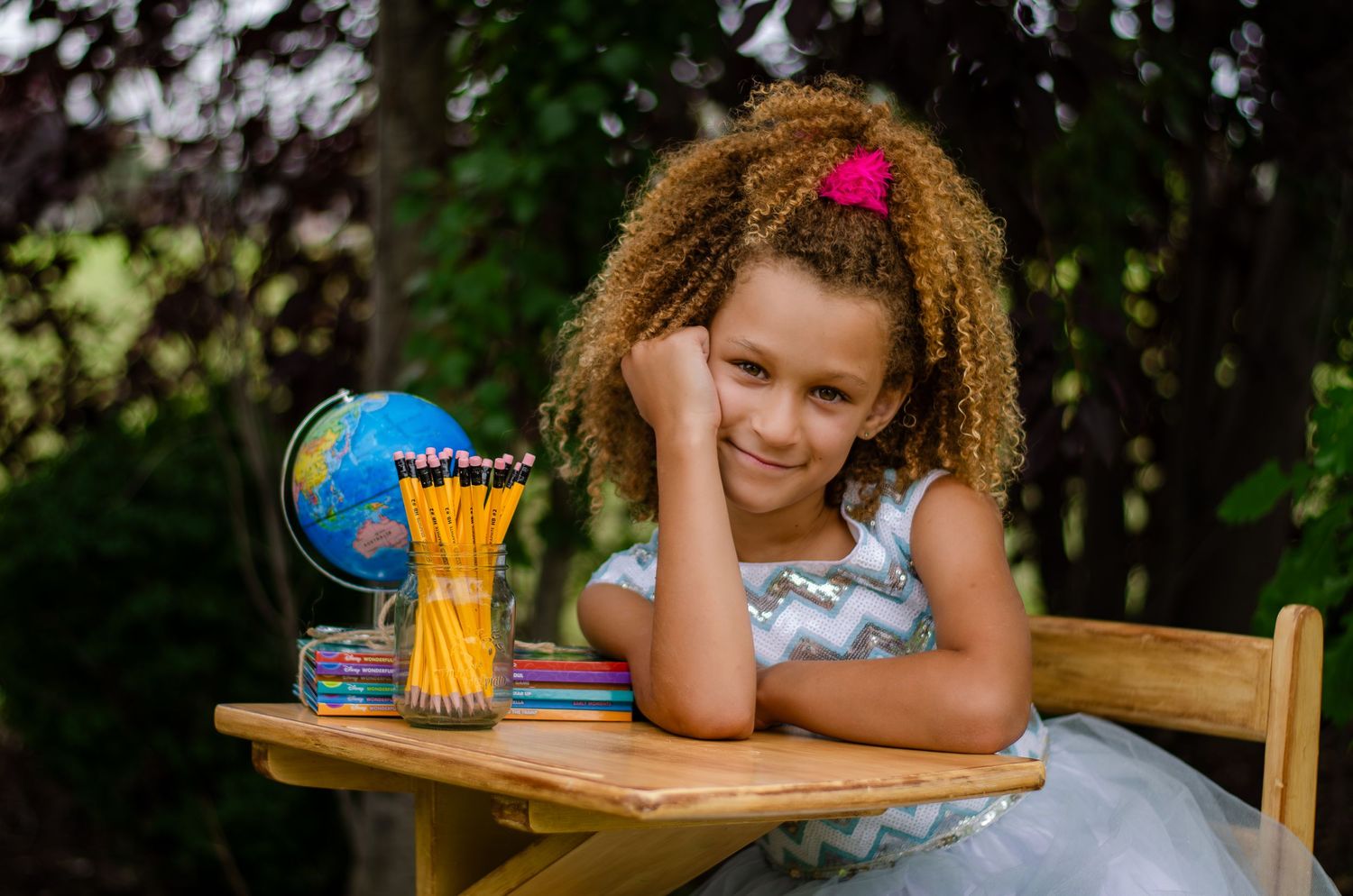 Student sits at wooden desk outdoors with globe and pencils, displaying a cheerful expression while learning.