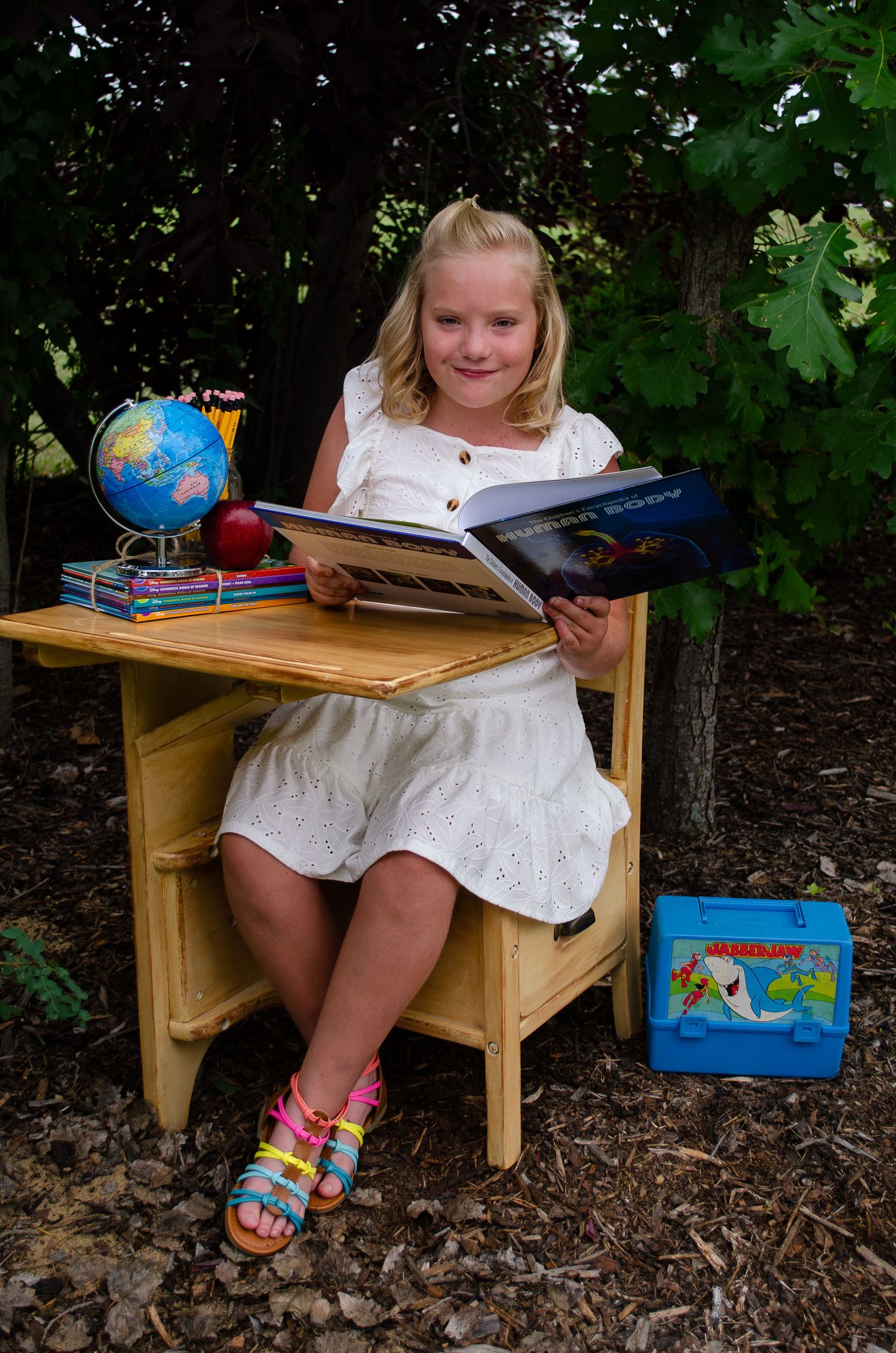 Child in white dress reading at wooden desk outdoors with globe and colorful lunchbox nearby.