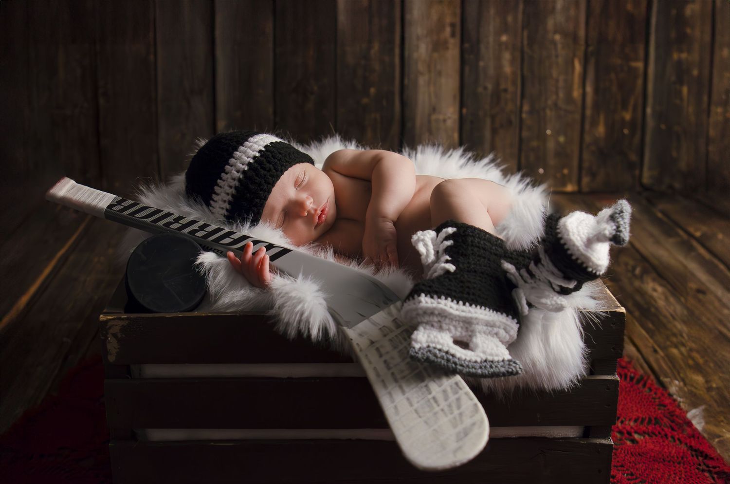 Sleeping newborn in hockey outfit with miniature stick and puck on wooden crate.