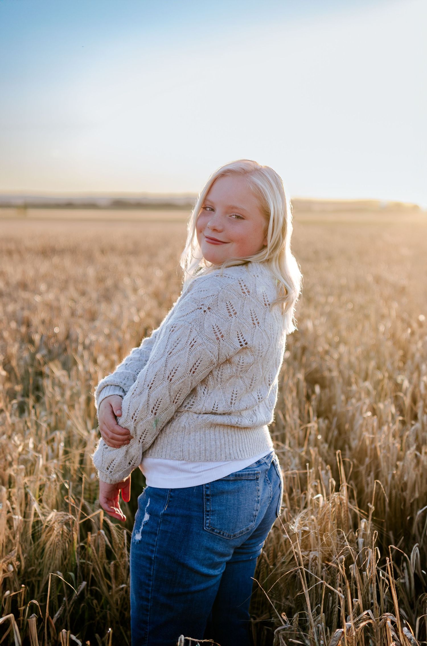 Woman in a white knit sweater poses in a golden wheat field at sunset.