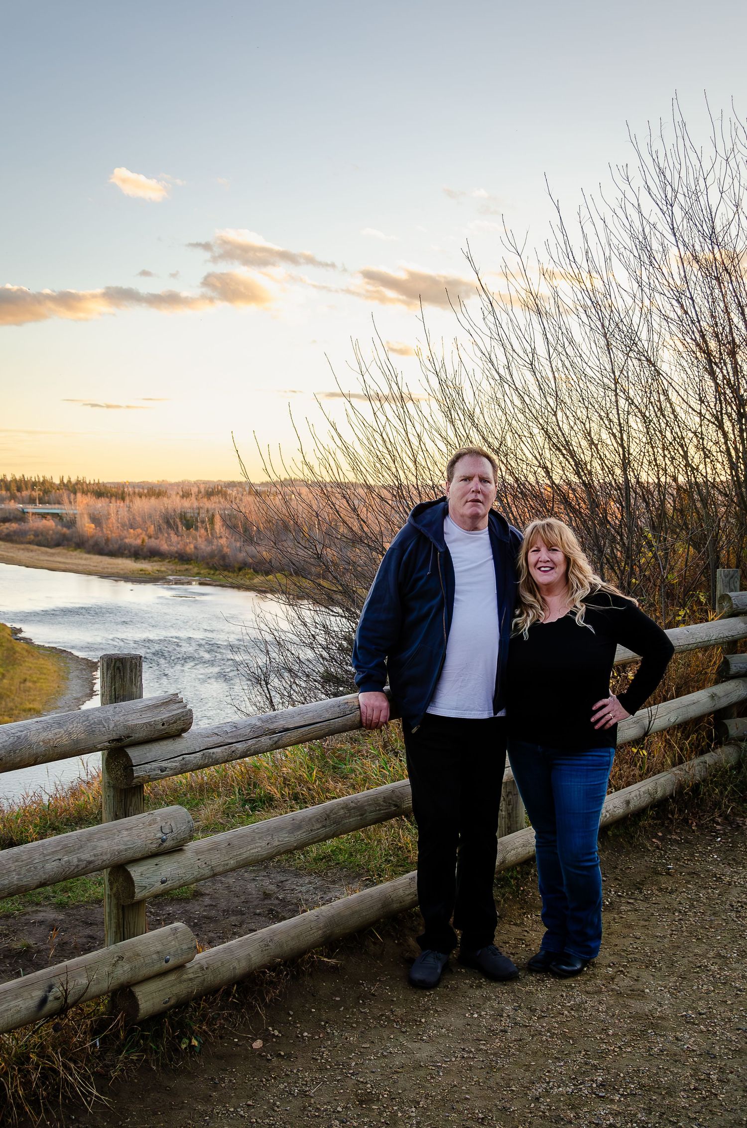 A couple stands together against a wooden fence at sunset overlooking a scenic river view.
