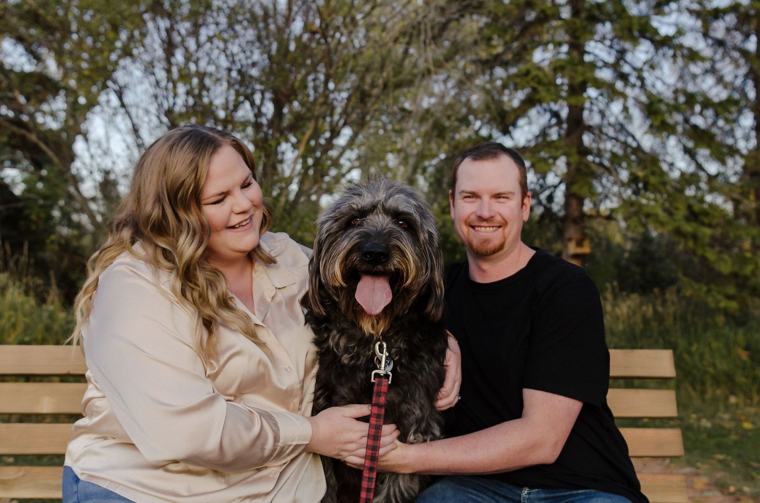A large black shaggy dog sits happily between two people on a wooden bench in an outdoor setting.