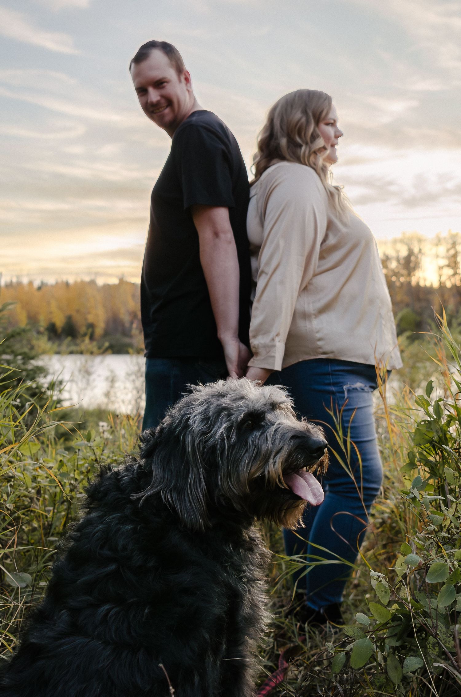 A large black shaggy dog stands with two people against a sunset backdrop by the water.