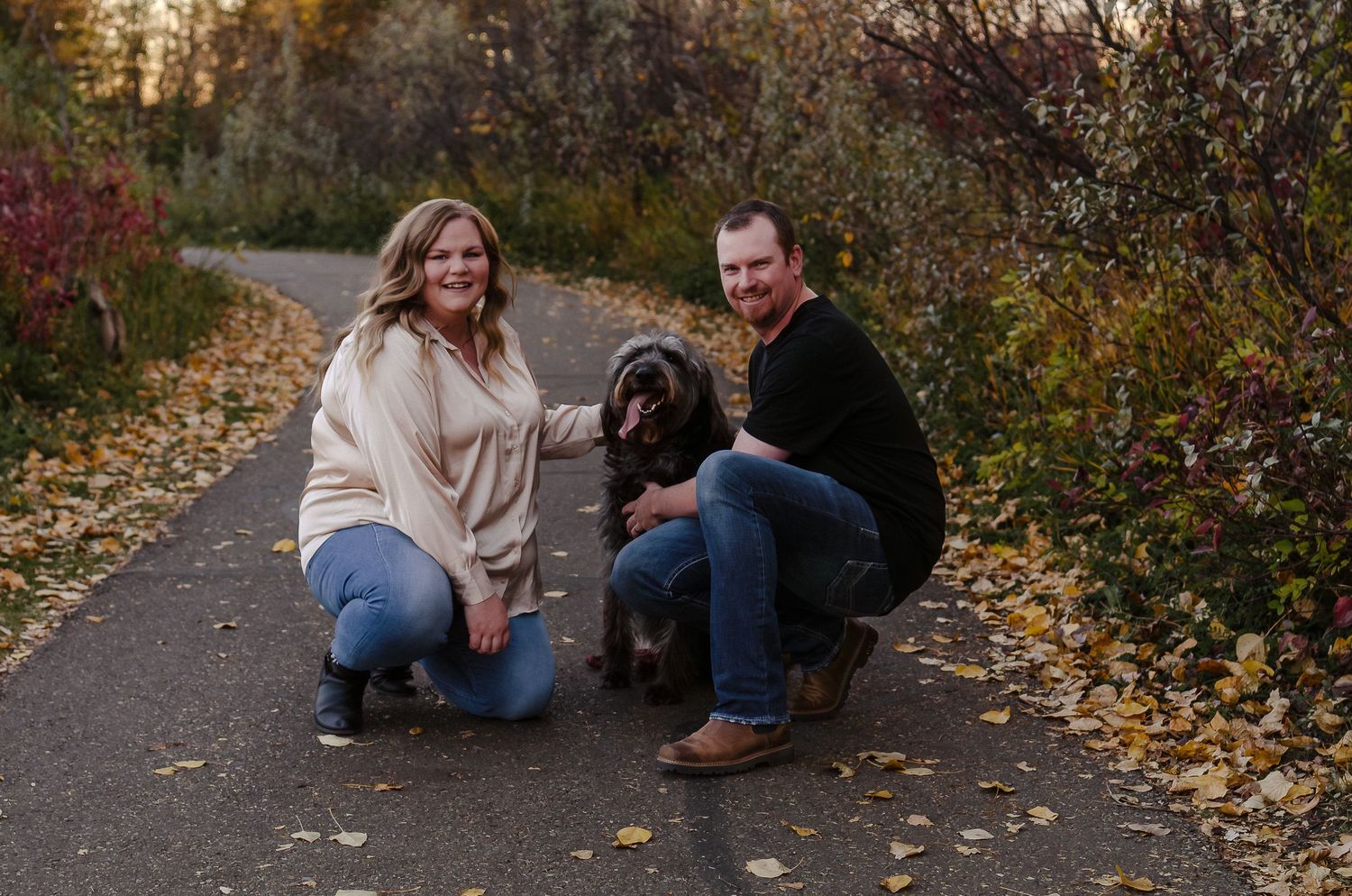 A couple crouches down with their black shaggy dog on an autumn path surrounded by fallen leaves.