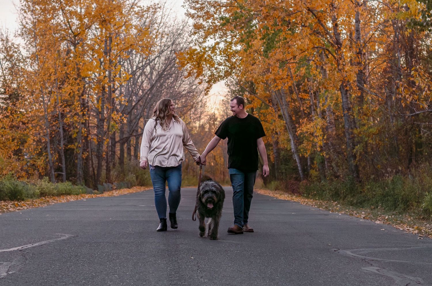 A couple walks their large black dog down a tree-lined path during autumn.