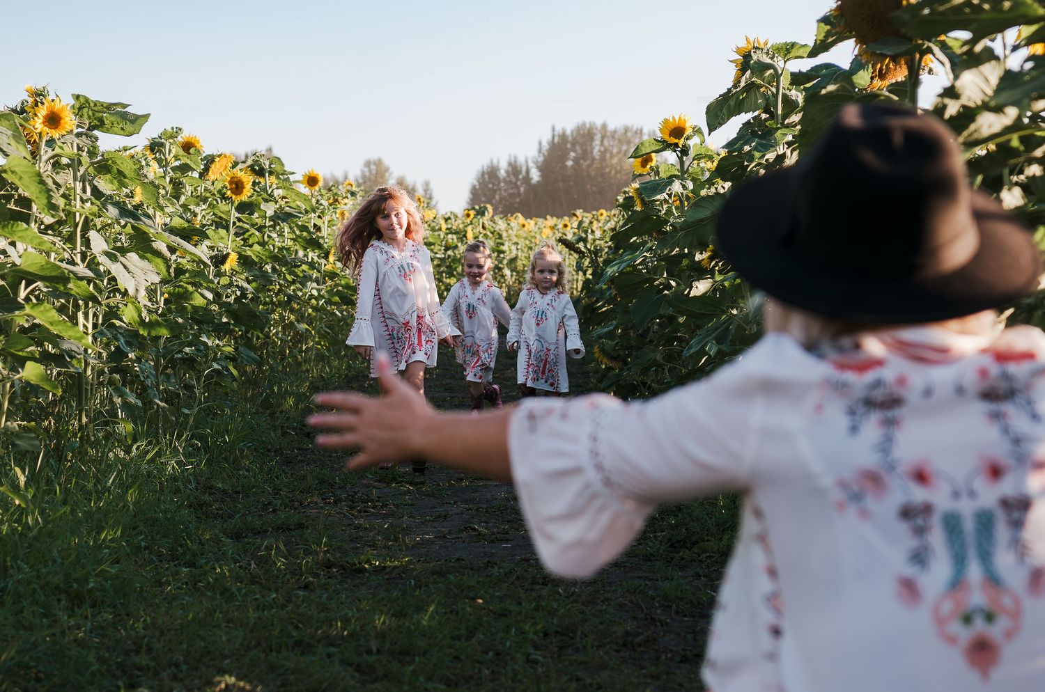 People in white floral dresses play together in a sunflower field path.