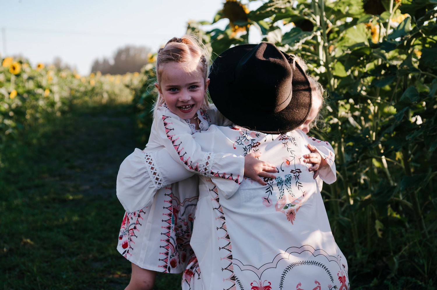 Two people in matching floral outfits embrace among tall sunflower stalks.