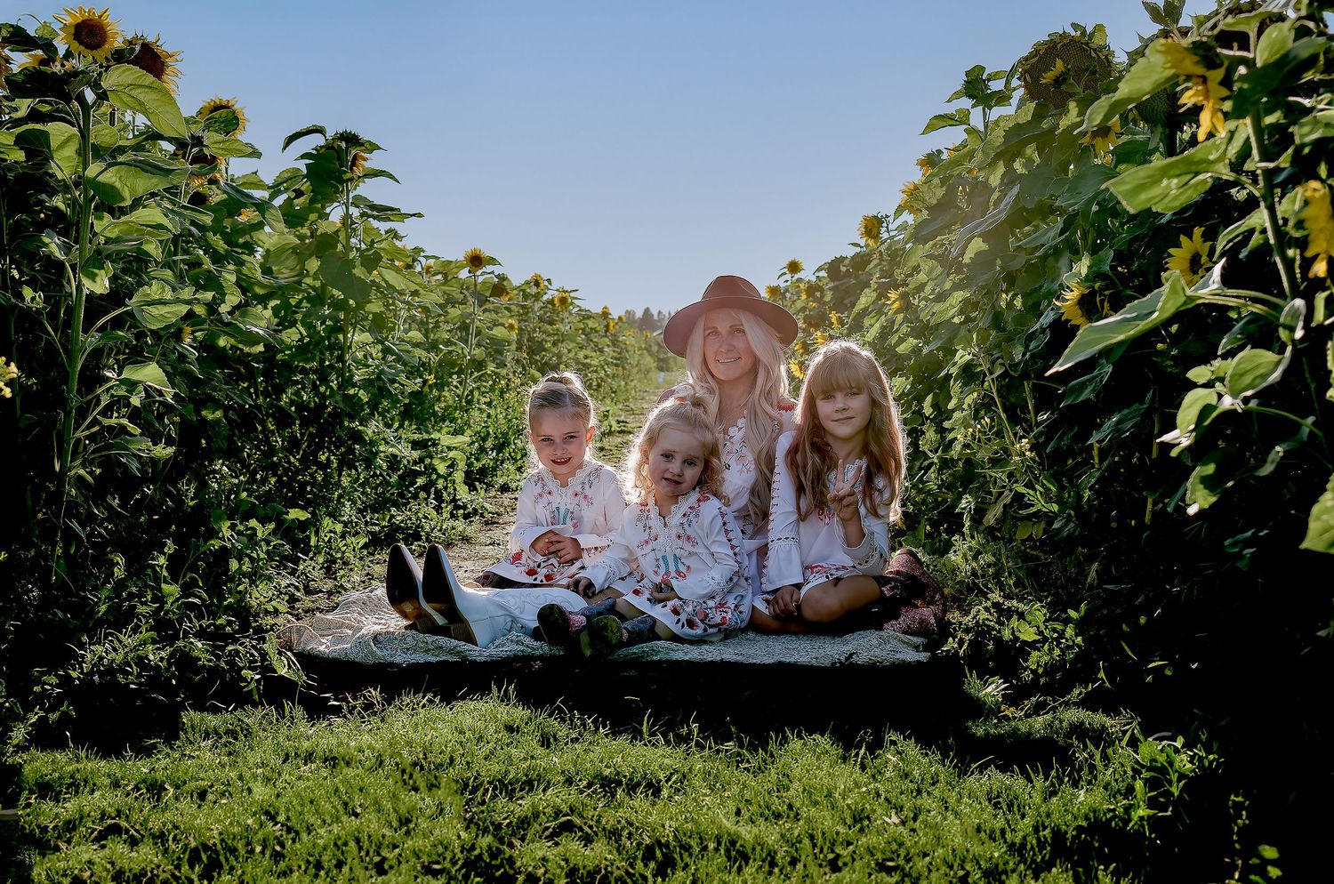 A group sits on a blanket between rows of tall sunflowers on a sunny day.