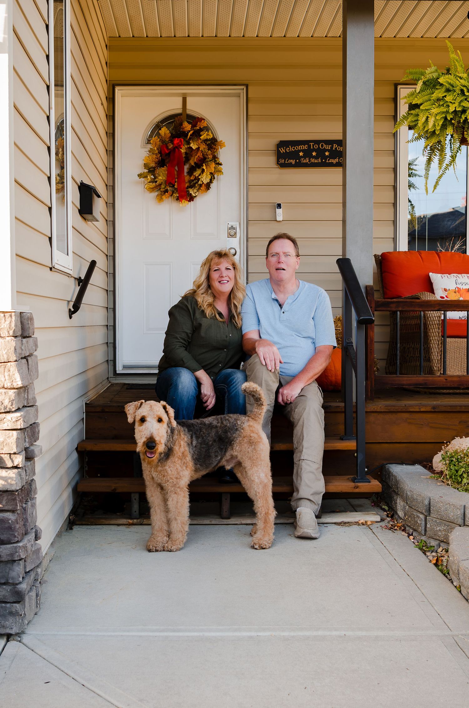 Couple sitting on front porch steps with their Airedale Terrier dog and autumn wreath on door.