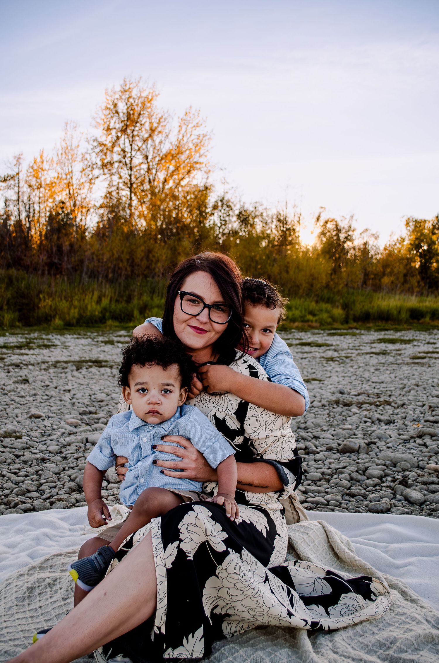 A family group sits together on a white blanket against a natural backdrop at sunset.
