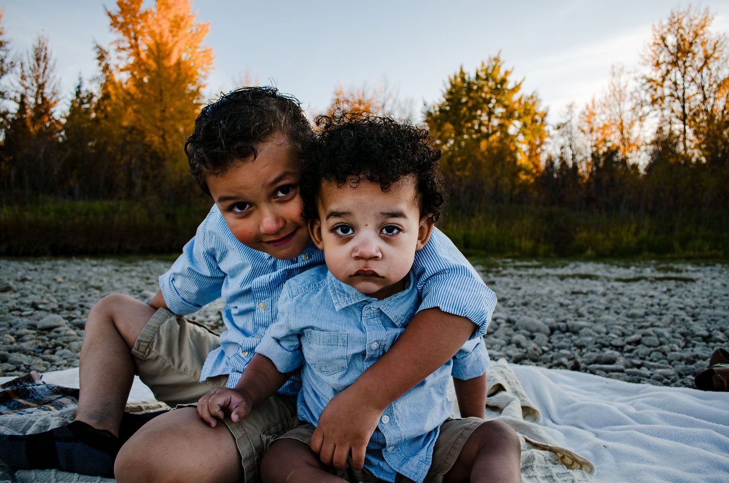 Two small children in matching blue shirts sit together on a white blanket outdoors.