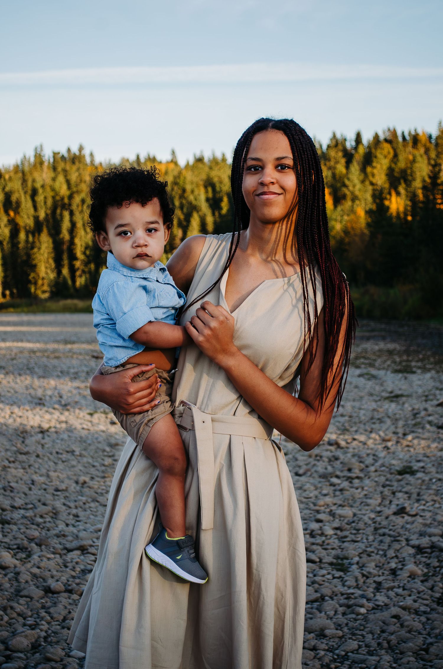 A person in a beige dress holds a small child wearing blue at sunset with evergreen trees in background.