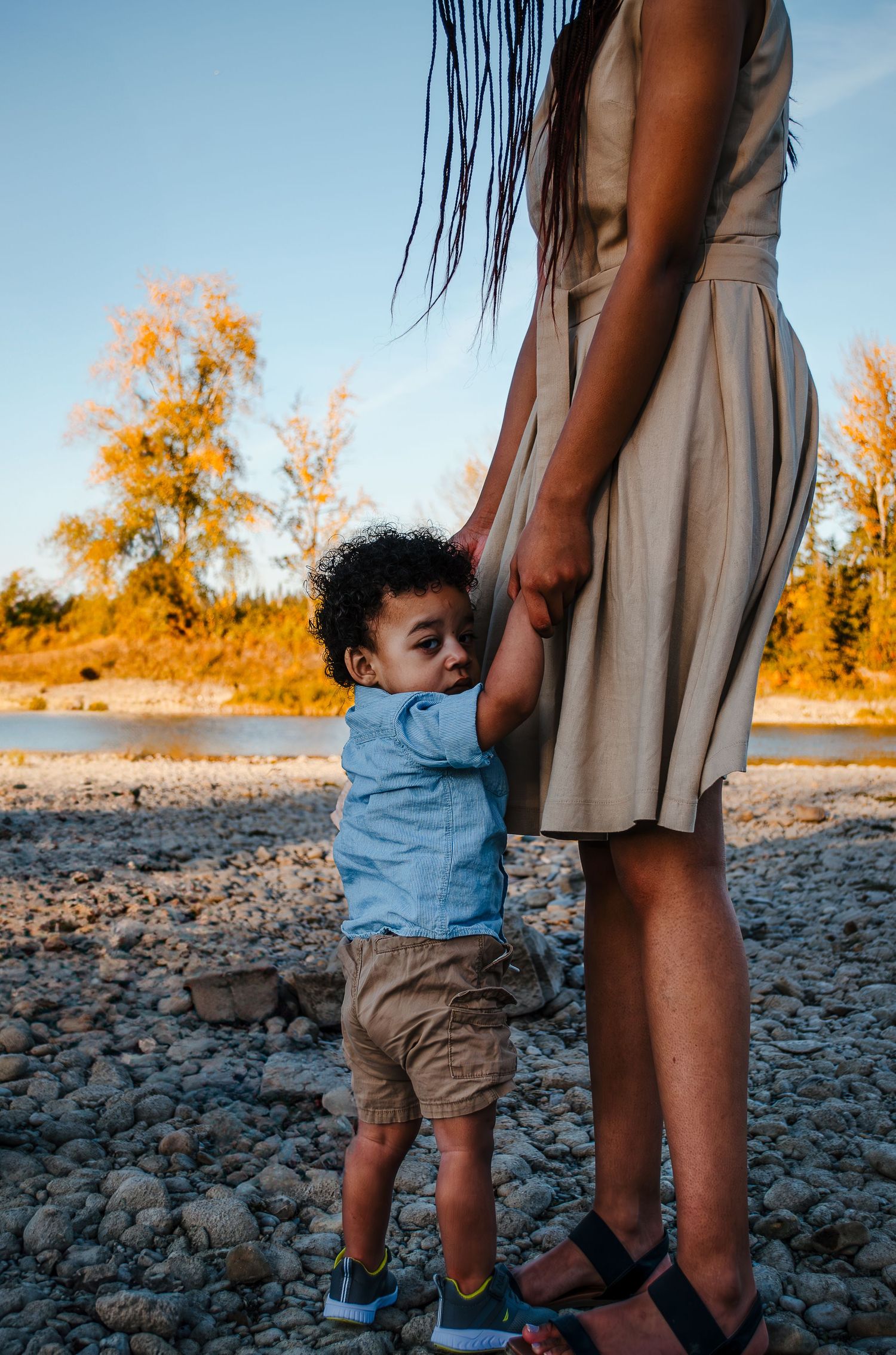 A small child in blue holds onto a person wearing a beige dress on a rocky beach at sunset.