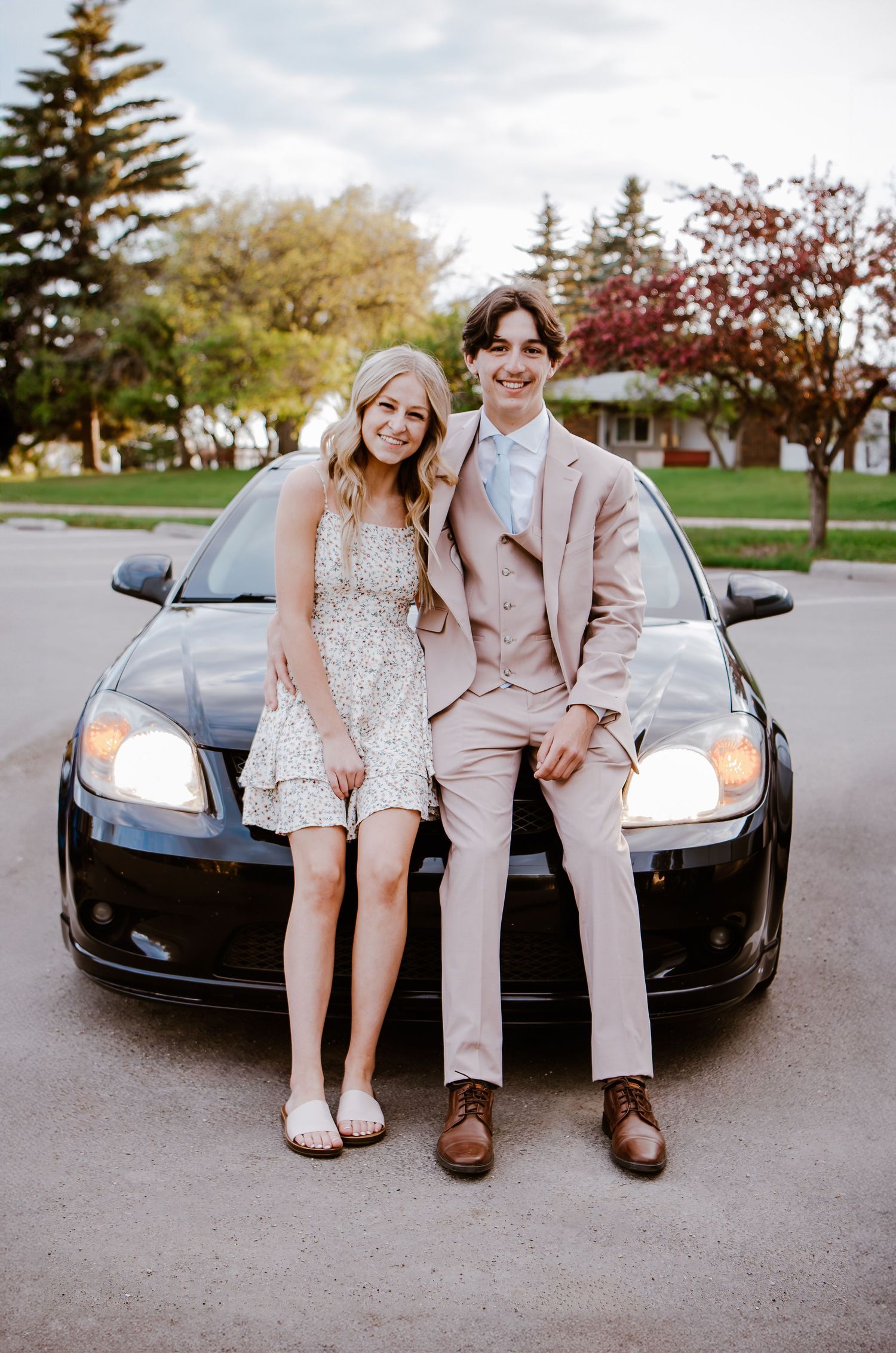 Couple in formal wear poses against black car with headlights on at dusk.