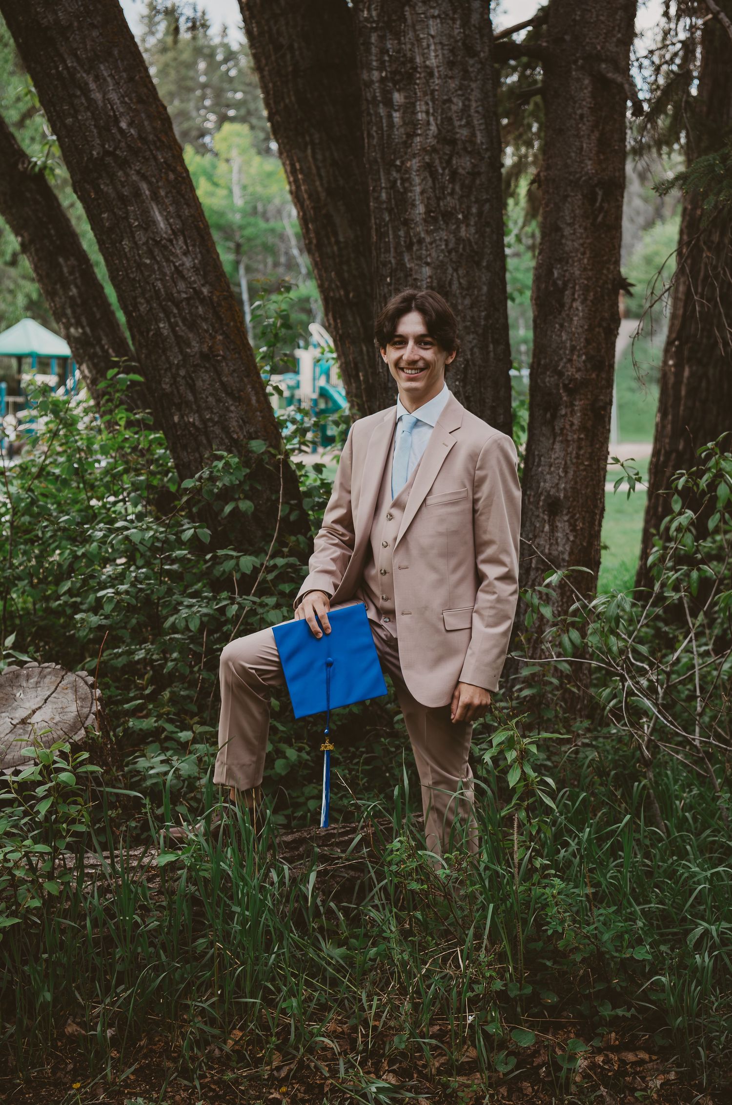 Person in beige suit poses with blue book among trees in woodland setting.