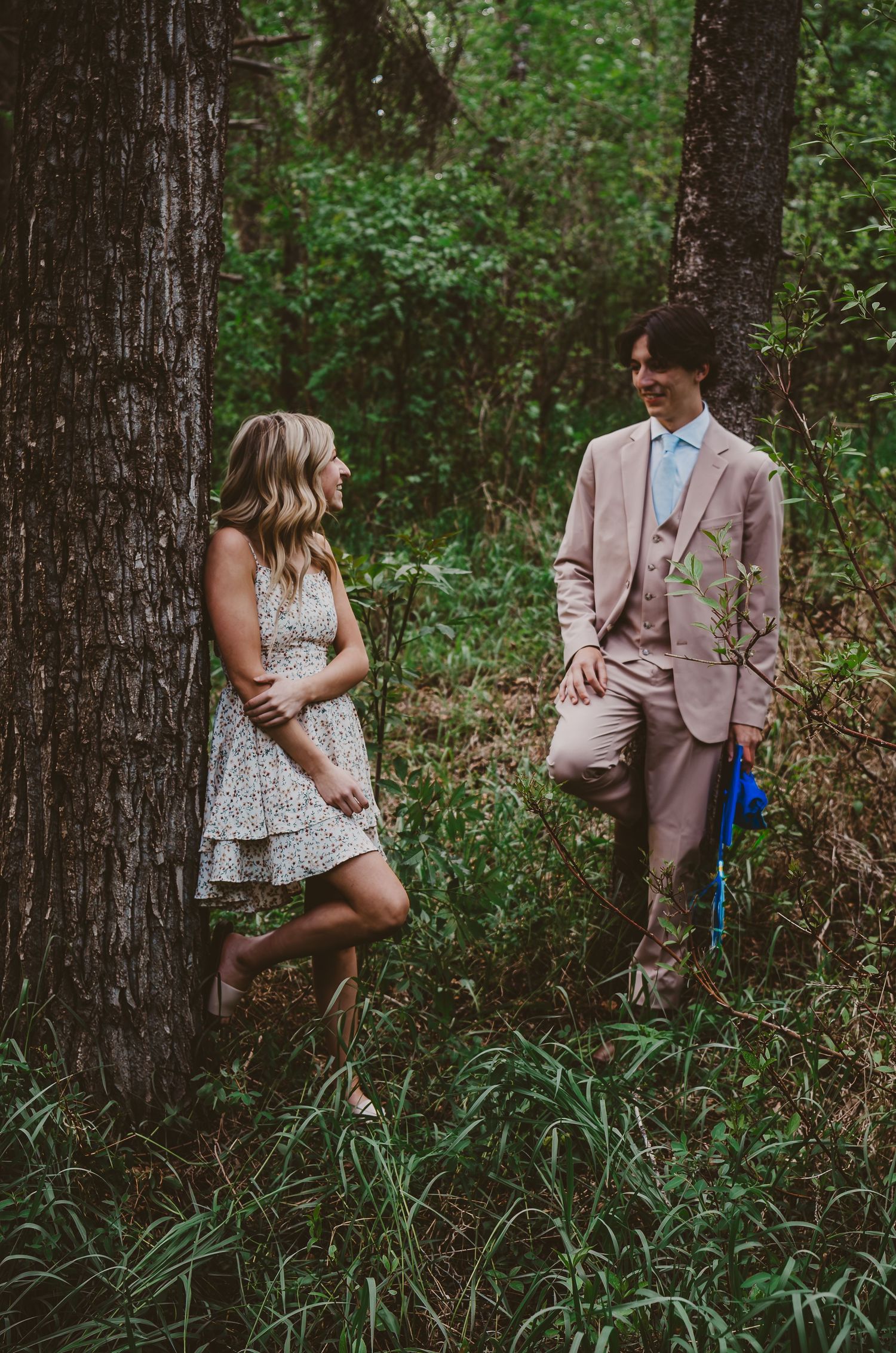 Couple in formal attire sharing a romantic moment while leaning against trees in a forest setting.