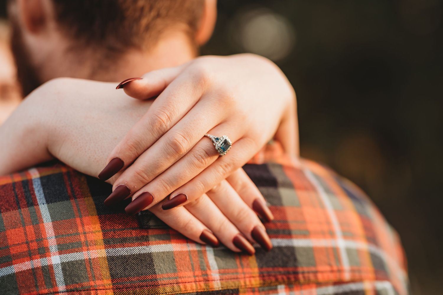 Close up of hands with dark nail polish and diamond engagement ring resting on plaid shirt in warm lighting.