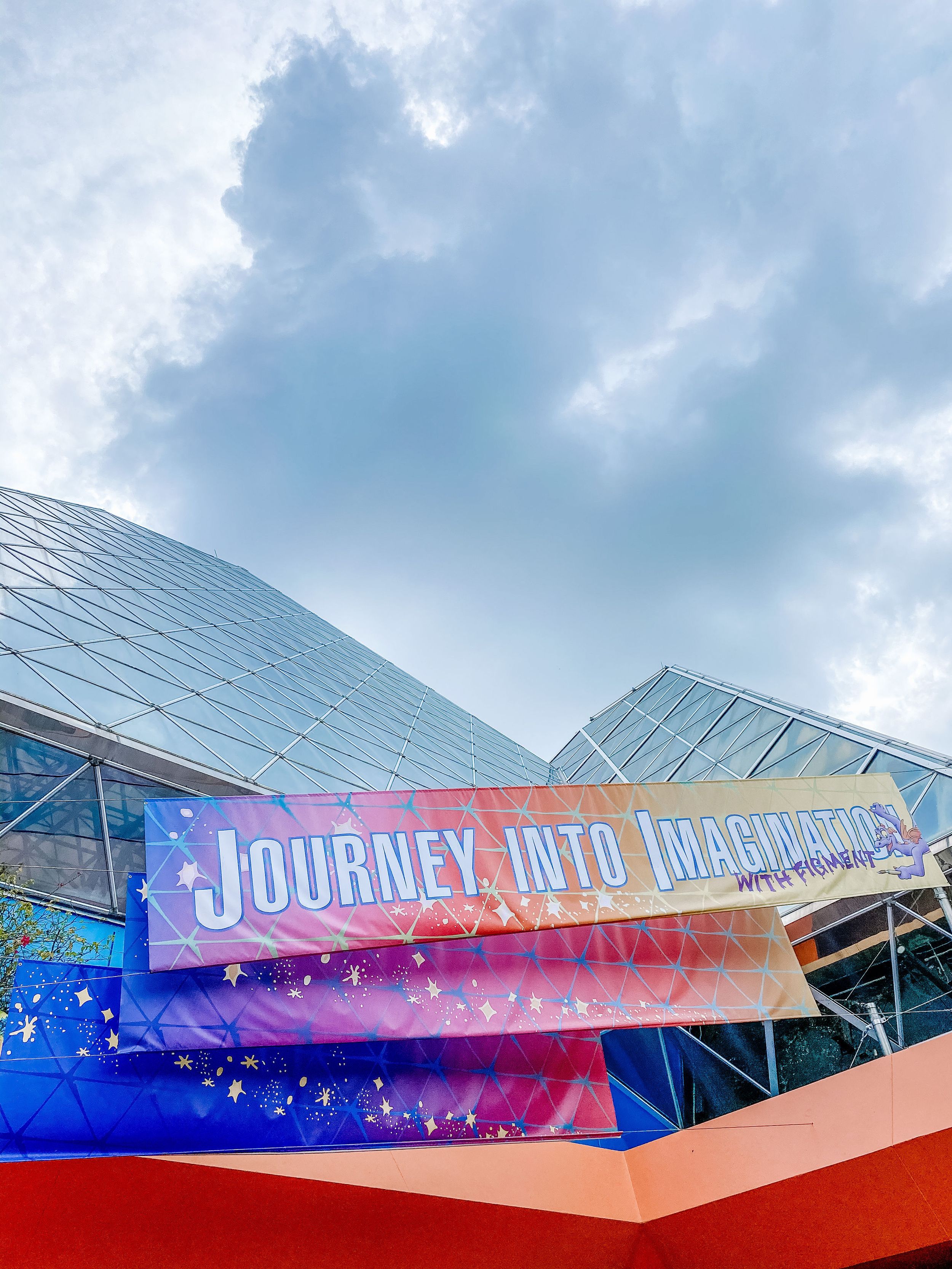 A Journey into Imagination sign displayed on a modern glass building facade under cloudy skies.