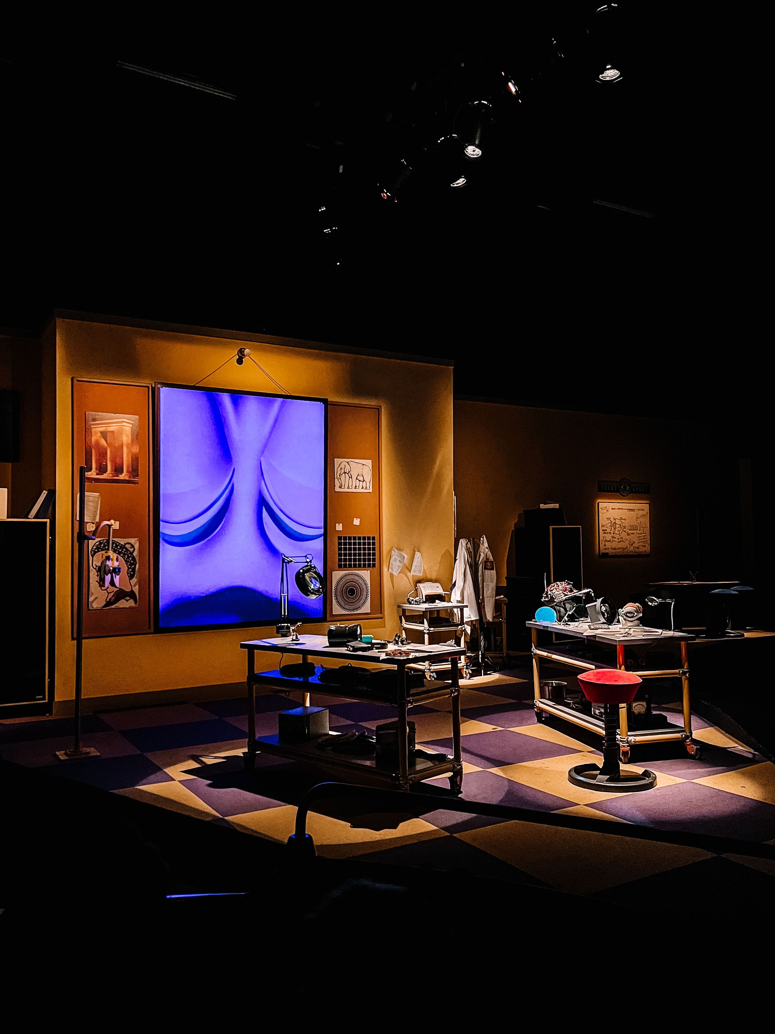 Dark theater stage setup with dramatic lighting, multiple furniture pieces, and orange-tinted walls creating an atmospheric mood.