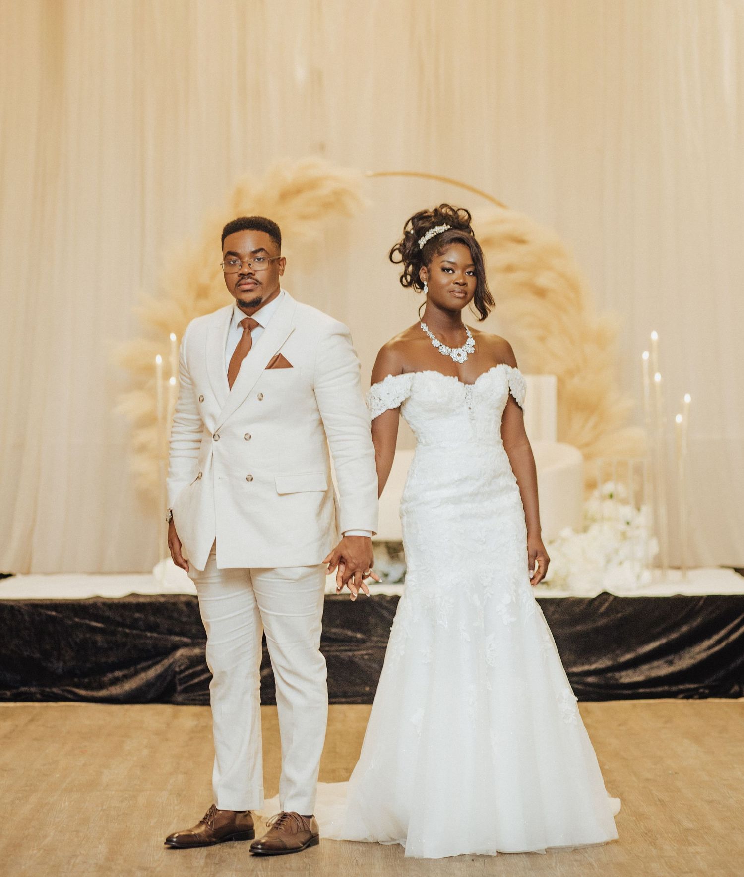 Elegant couple in coordinated white wedding attire holding hands in front of a softly lit decorative backdrop.