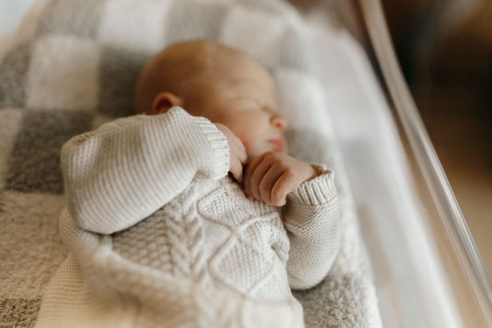 Newborn baby wearing cozy white knit sweater and hat while sleeping on gray blanket.