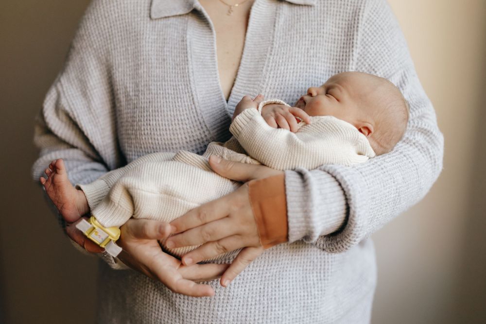 Baby in white outfit being held while sleeping in caregiver's gray sweater.