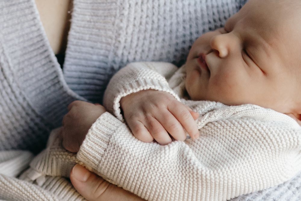 Sleeping baby in white knit sweater rests peacefully against gray fabric.