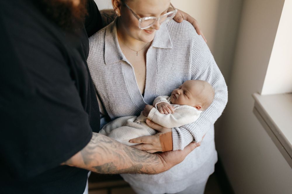 A tender moment as a couple cradles their newborn baby while standing near a window in natural light.