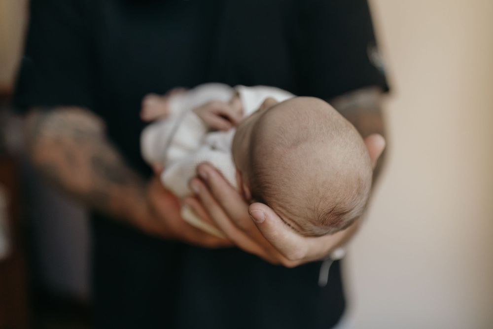 Close-up view of tattooed arms holding a sleeping newborn baby wrapped in white.