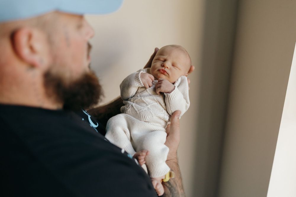 A person wearing a blue cap tenderly holds a sleeping newborn dressed in white.