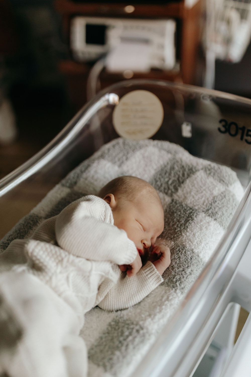 Sleeping newborn in white sweater rests peacefully in hospital bassinet.