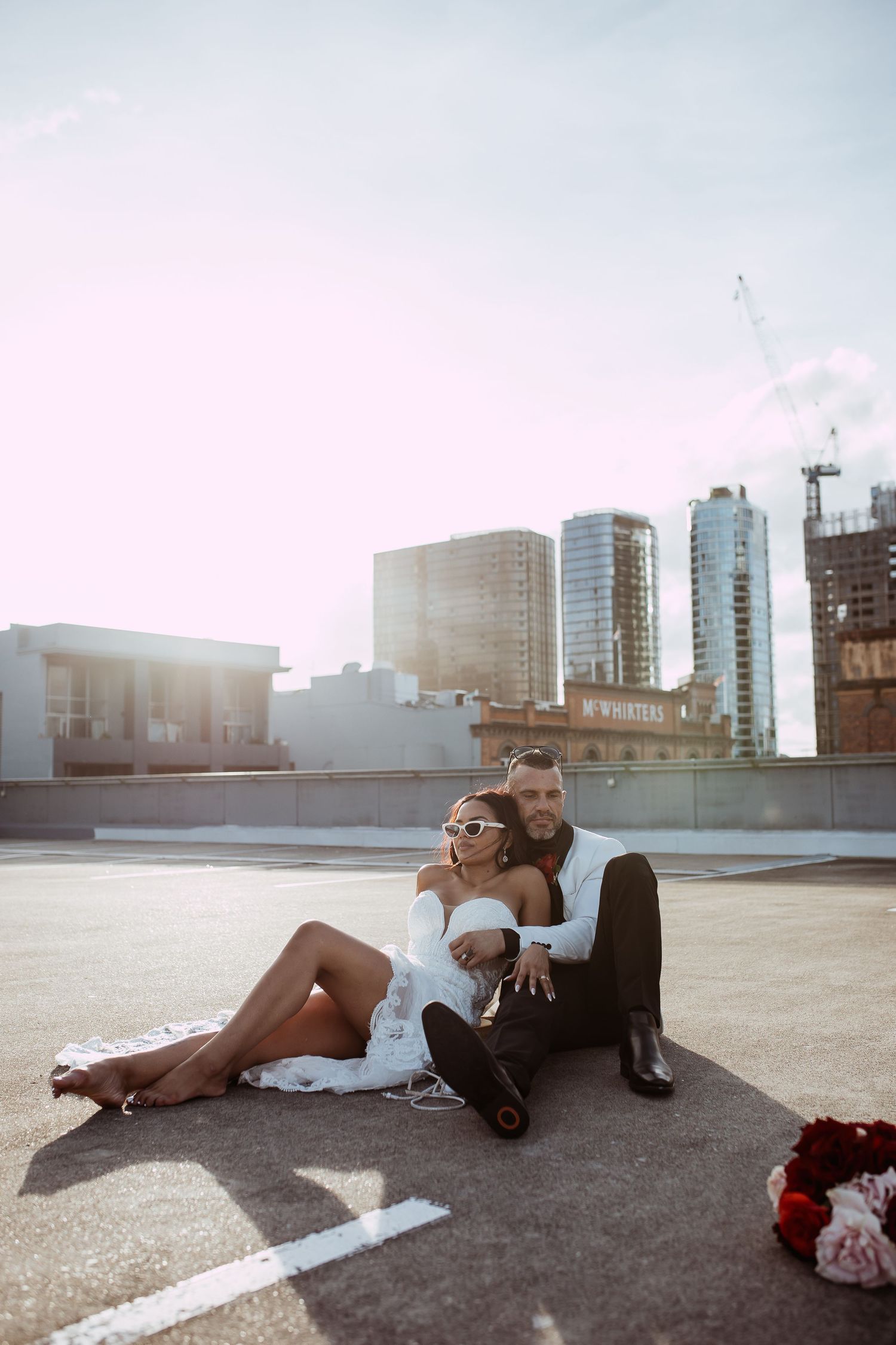 A stylish couple sits together on a rooftop at sunset with city buildings in the background.