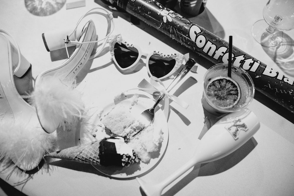 Black and white wedding flat lay with heart sunglasses, confetti cannon, heels, and festive accessories on white surface.