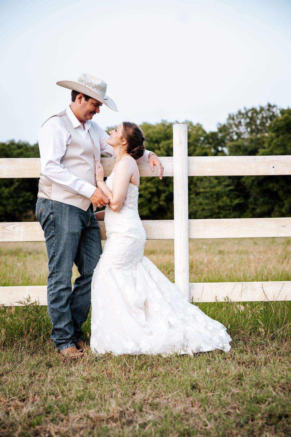 Bride and groom share a romantic moment by a white fence in a rustic outdoor setting, groom wearing cowboy hat.
