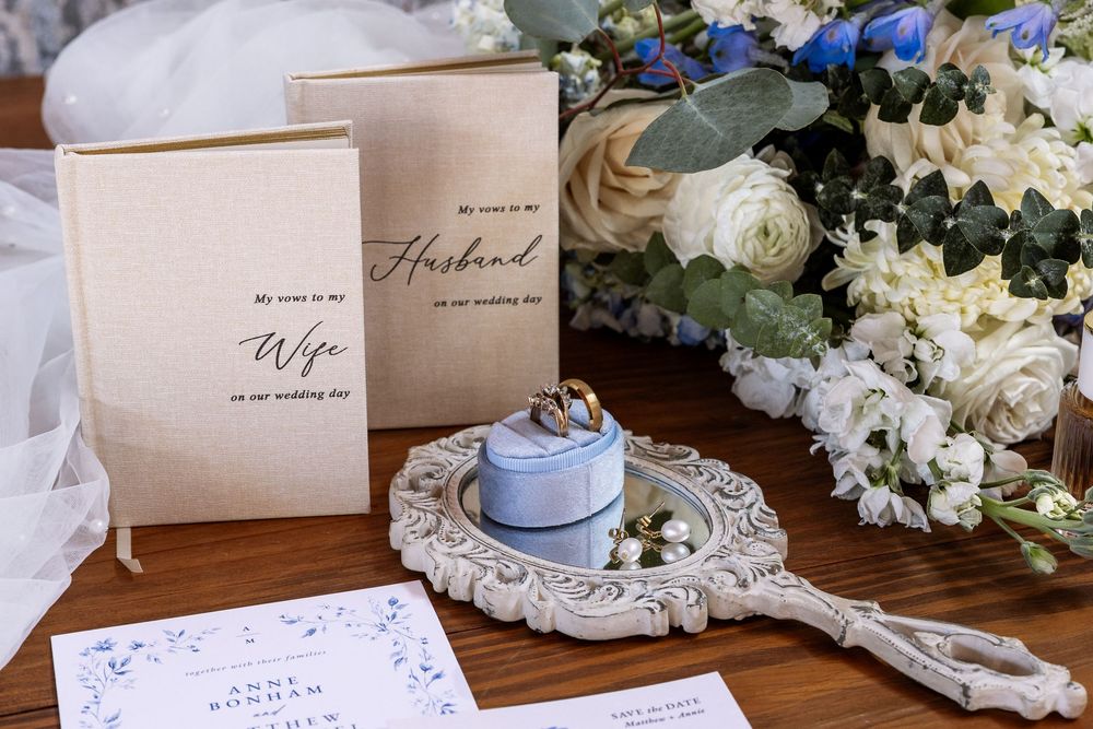 Wedding detail flat lay with vow books, blue ring box, wedding rings, floral bouquet, and stationery on wood table.