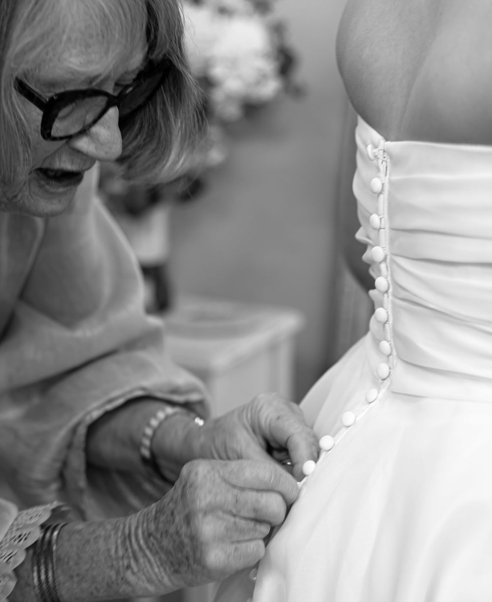 Black and white photo of elderly woman carefully buttoning the back of a bride's white wedding gown.