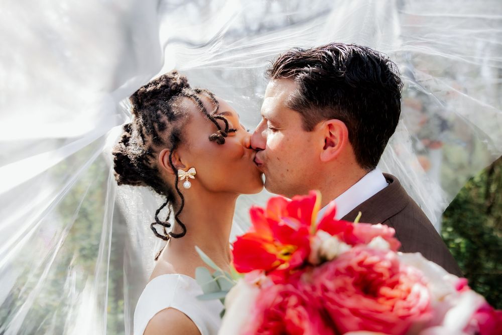 Bride and groom share a kiss under a flowing veil with a vibrant pink bouquet in the foreground.