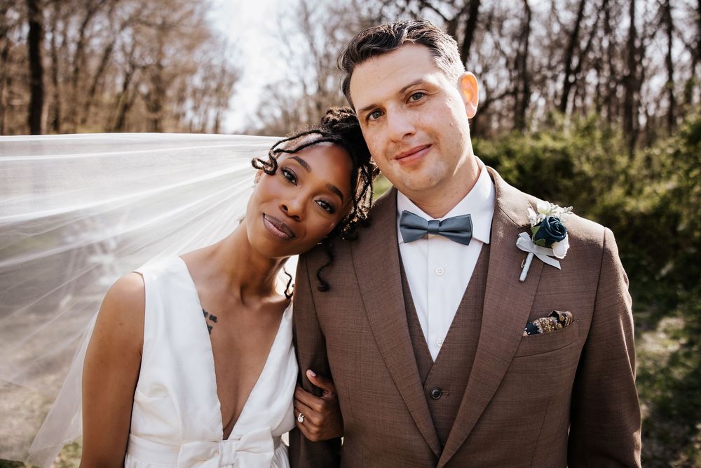 Interracial couple on their wedding day, bride leaning on groom in brown suit with boutonniere.